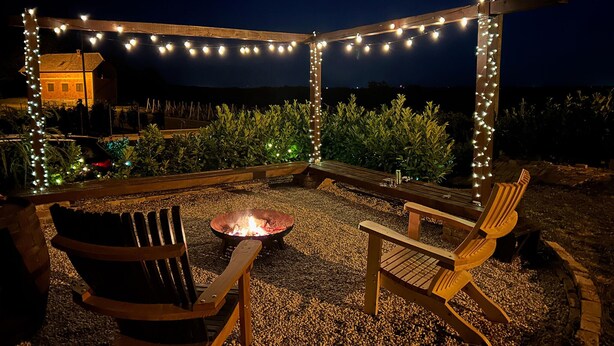 An outdoor seating area with wooden chairs and a fire pit, surrounded by string lights and gravel.