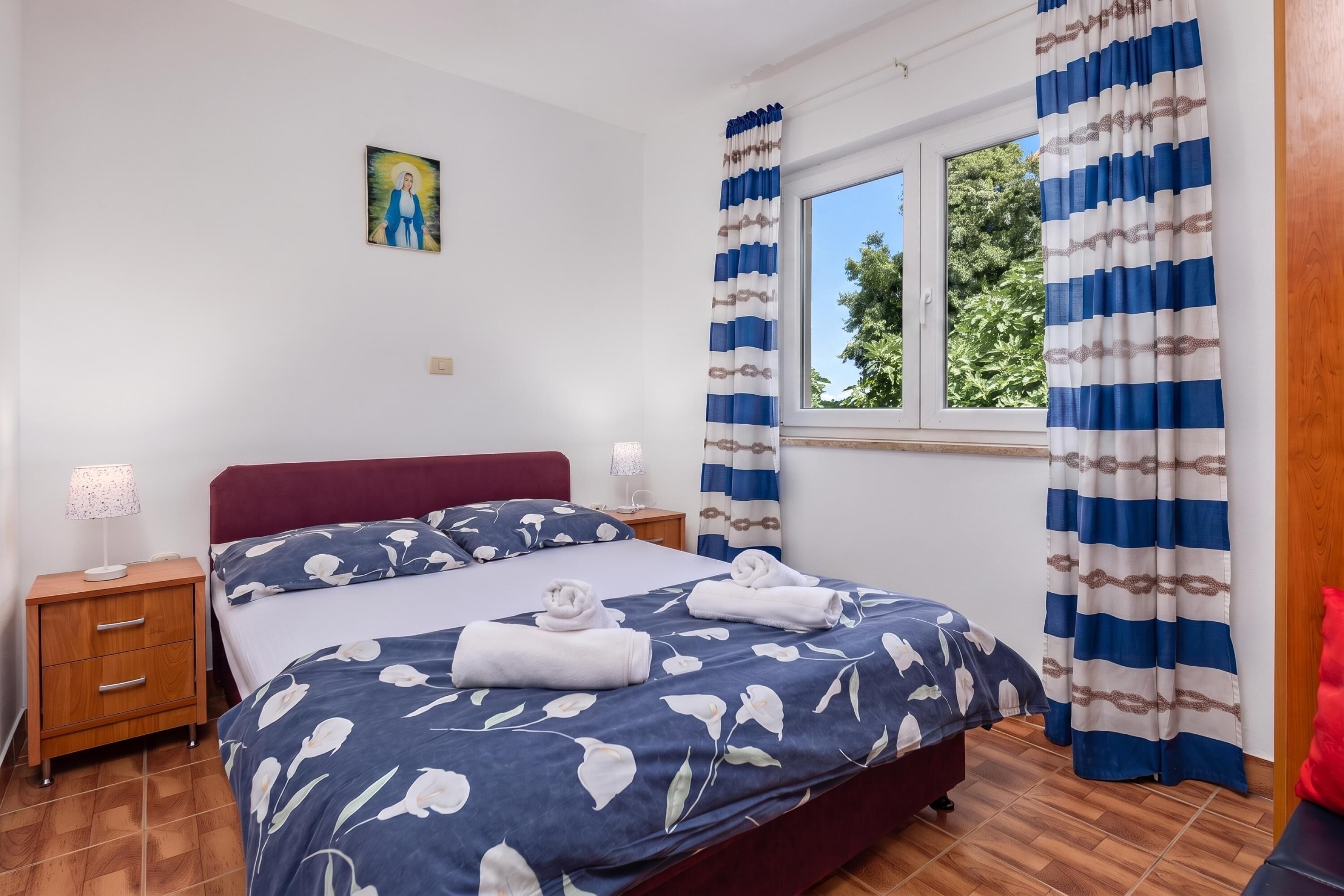 Quarto de hotel com cama, mesa de cabeceira, luminária, janela com cortinas e um quadro na parede.