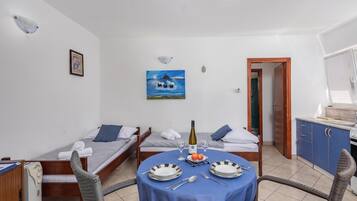 Una habitación de hotel compacta con dos camas individuales, una mesa de comedor con sillas, una pequeña cocina y un cuadro en la pared.