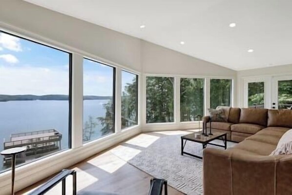 A spacious living room with a large window overlooking a lake, a brown leather sofa, and a small table.
