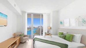 A modern hotel room with a large bed, a desk, a chair, a view of the city and water through large windows. - Amazing 1BR Apartment + Pool + Fitness Center (Hallandale Beach)