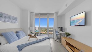 A modern hotel room with a large bed, a desk, and a view of the cityscape through the window. - Spectacular 1BR Apt w/Private Balcony, pool & Gym (Hallandale Beach)