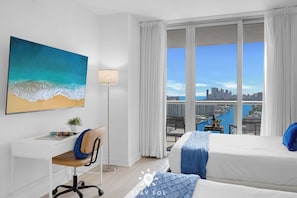 A hotel room with a city view, a desk with a chair, a bed with blue bedding, and a painting of a beach.