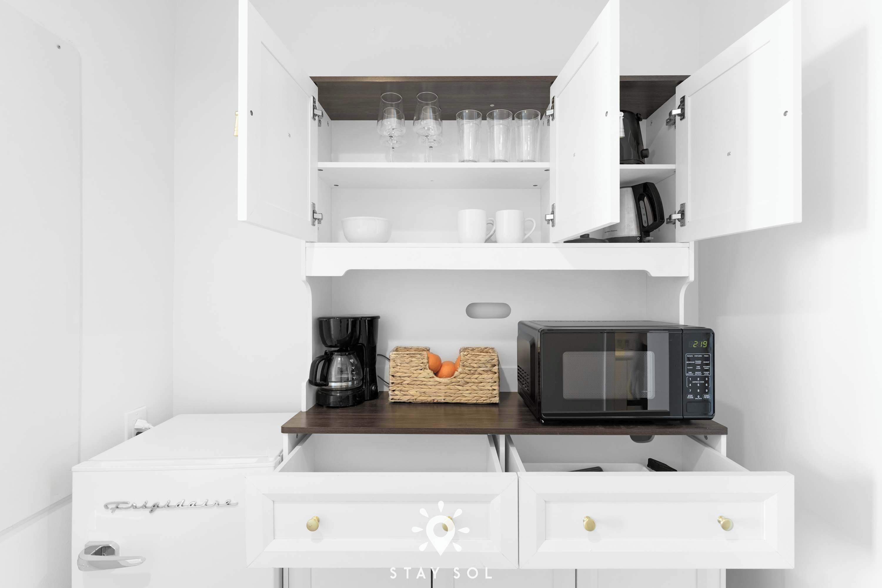 Een kleine keuken met witte kastjes, een magnetron, een koffiezetapparaat en een rieten mandje op het aanrecht.