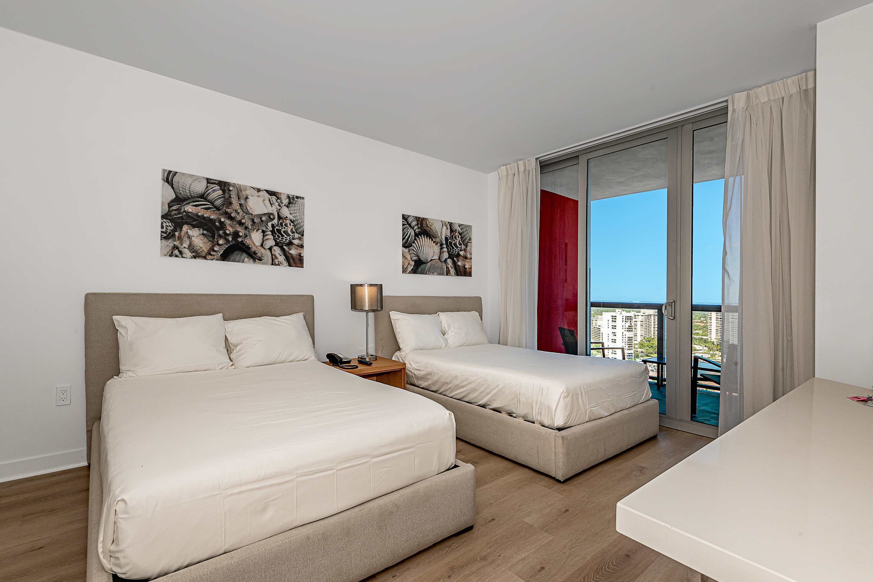 A hotel room with two beds, a desk, and a balcony with a city view.