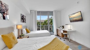 Habitación de hotel con dos camas, un escritorio, una silla y un amplio ventanal con vista a palmeras.