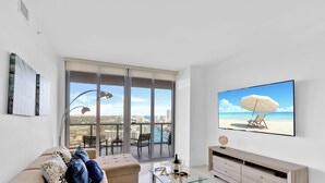 A modern living room with a large window, a sofa, a TV, and a view of a beach. - Stylish - Incredible Views & Top Amenities (Hallandale Beach)