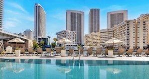 Una piscina con sillones de descanso, un área de estar techada y edificios altos al fondo.