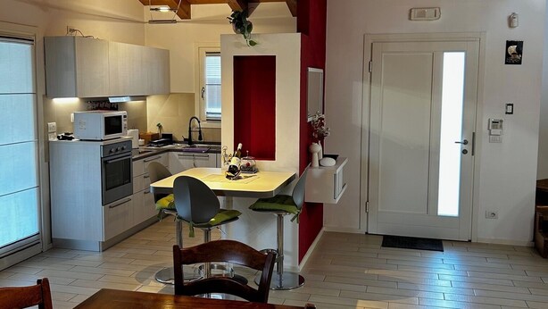 A modern kitchen with a dining area, wooden beams on the ceiling, and a red accent wall. - A cozy and serene place for those seeking tranquility and comfort (Porpetto)