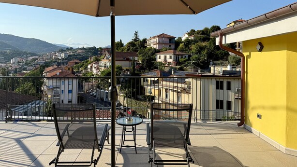 Una terraza con una mesa, sillas, un paraguas grande y vistas a un pueblo en la ladera.