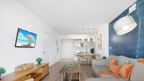 Ein modernes Wohnzimmer mit Sofa, Couchtisch und Fernseher. Im Hintergrund ist eine Küche zu sehen.