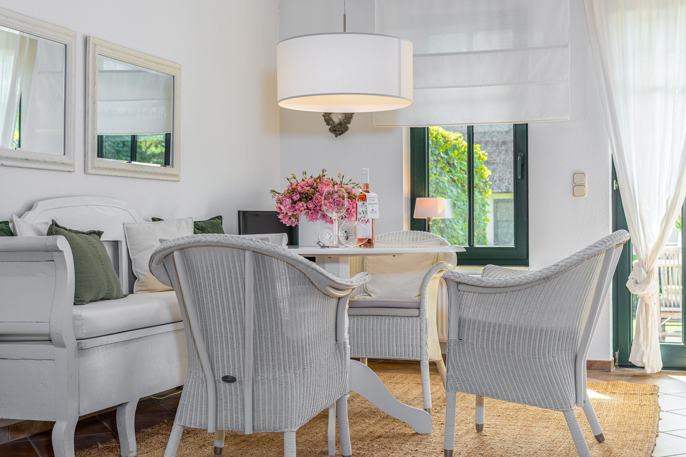 A modern living room with a white sofa, two wicker chairs, a dining table with a white lamp, and a large window with green shutters.