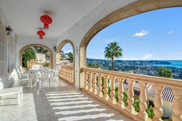A balcony with white chairs and tables, a railing, and a view of a coastal city.