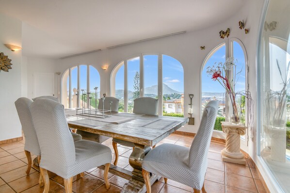 A dining area with a large table and chairs, large arched windows, and a view of a mountain.