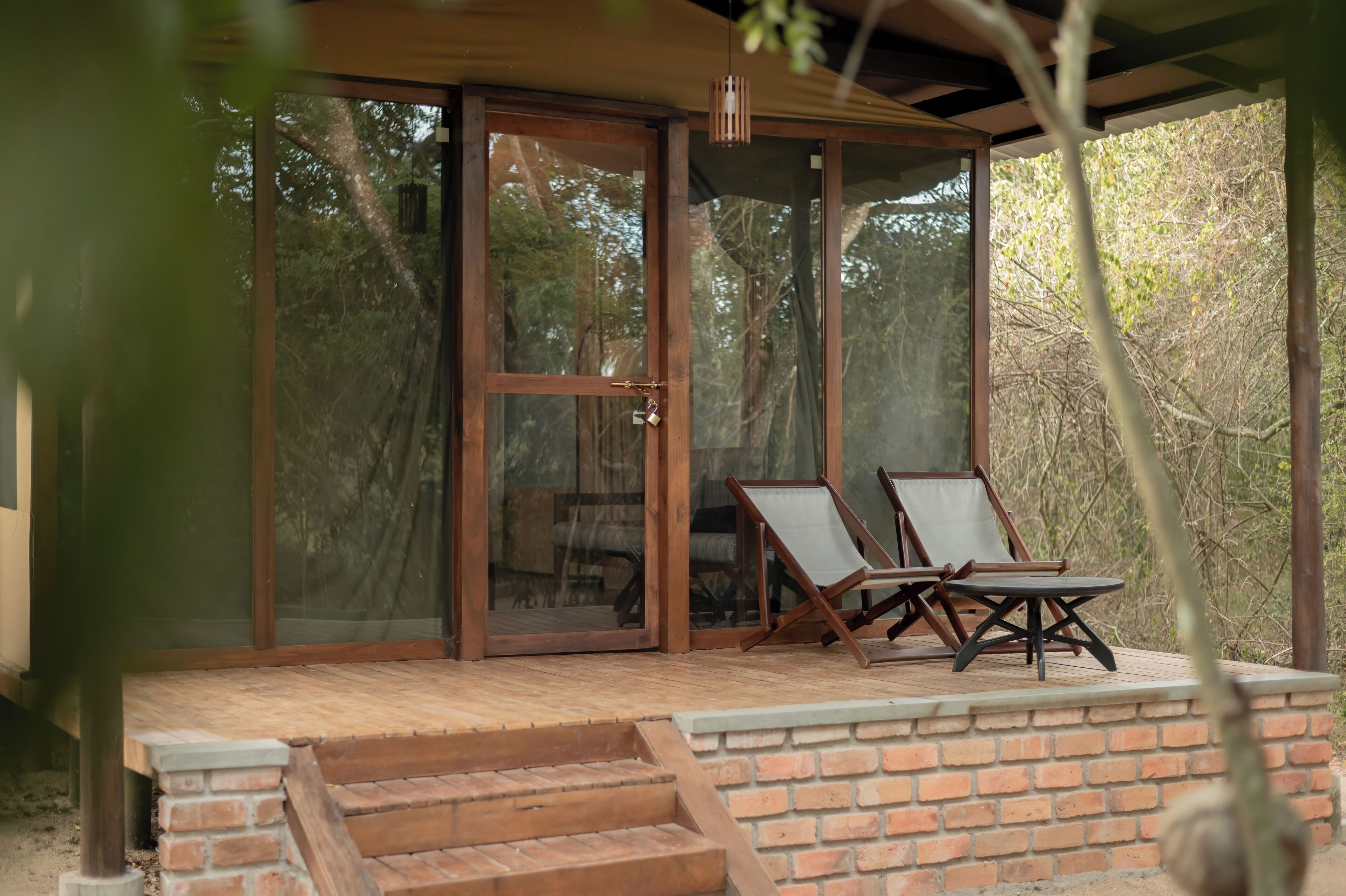 A covered outdoor seating area with two chairs and a table, a wooden door, and a brick wall.