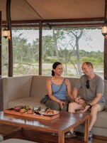 Una pareja sentada en un sofá en una habitación con una mesa de madera al frente, una ventana con vista a árboles, y una lámpara colgando del techo.
