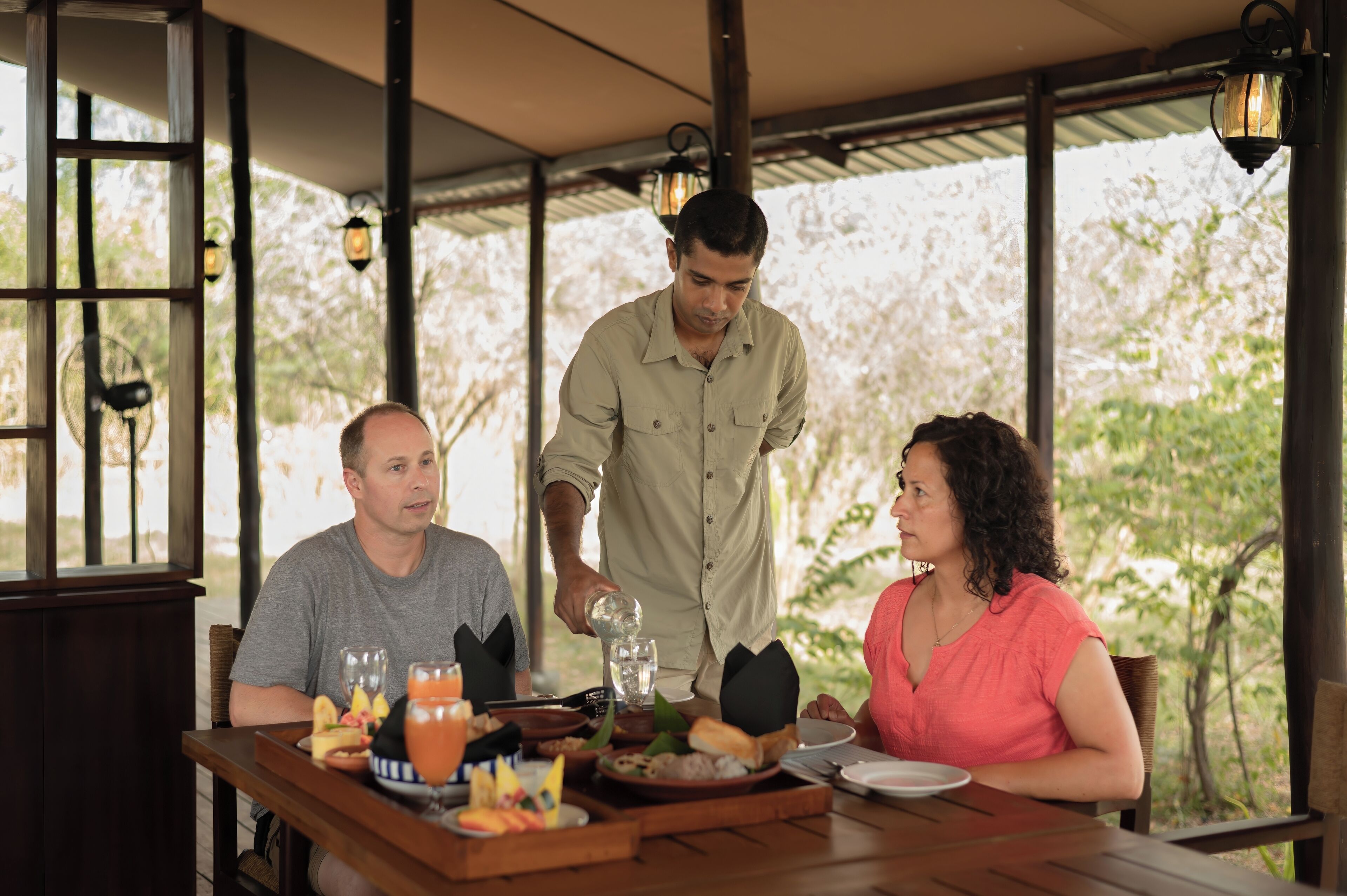 Uma área de jantar com uma mesa posta para três pessoas, um garçom servindo uma bebida e vista para árvores através de grandes janelas.