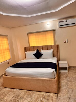 A hotel room with a large bed, a bedside table, an air conditioning unit, and a window with blinds. - Opal Selfie Apartment in Lagos (Lagos)
