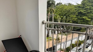 A balcony with a black wicker lounge chair, a metal railing, and a view of parked cars and greenery.