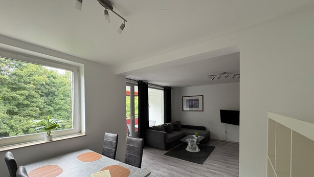 Een moderne woonkamer met een grijze bank, een salontafel en een televisie aan de muur. Er is een eethoek met een tafel en stoelen.