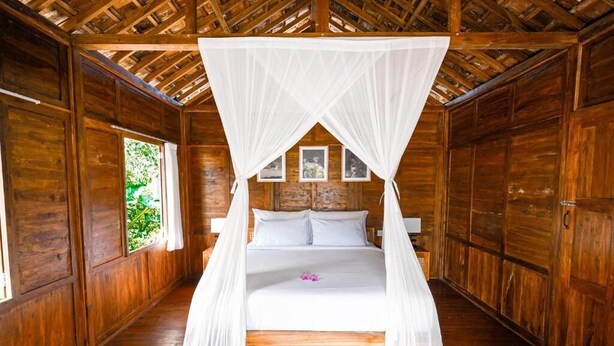 Una habitación de madera con una cama bajo un dosel blanco, paredes de madera y una ventana con follaje en el exterior.