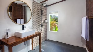 Un baño con tocador de madera, un espejo redondo, ducha con puerta de vidrio y ventana con vista a la vegetación.