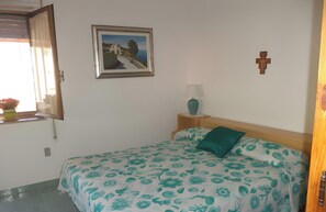 A bedroom with a bed, a window, a picture on the wall, a cross, and a bedside table with a lamp.