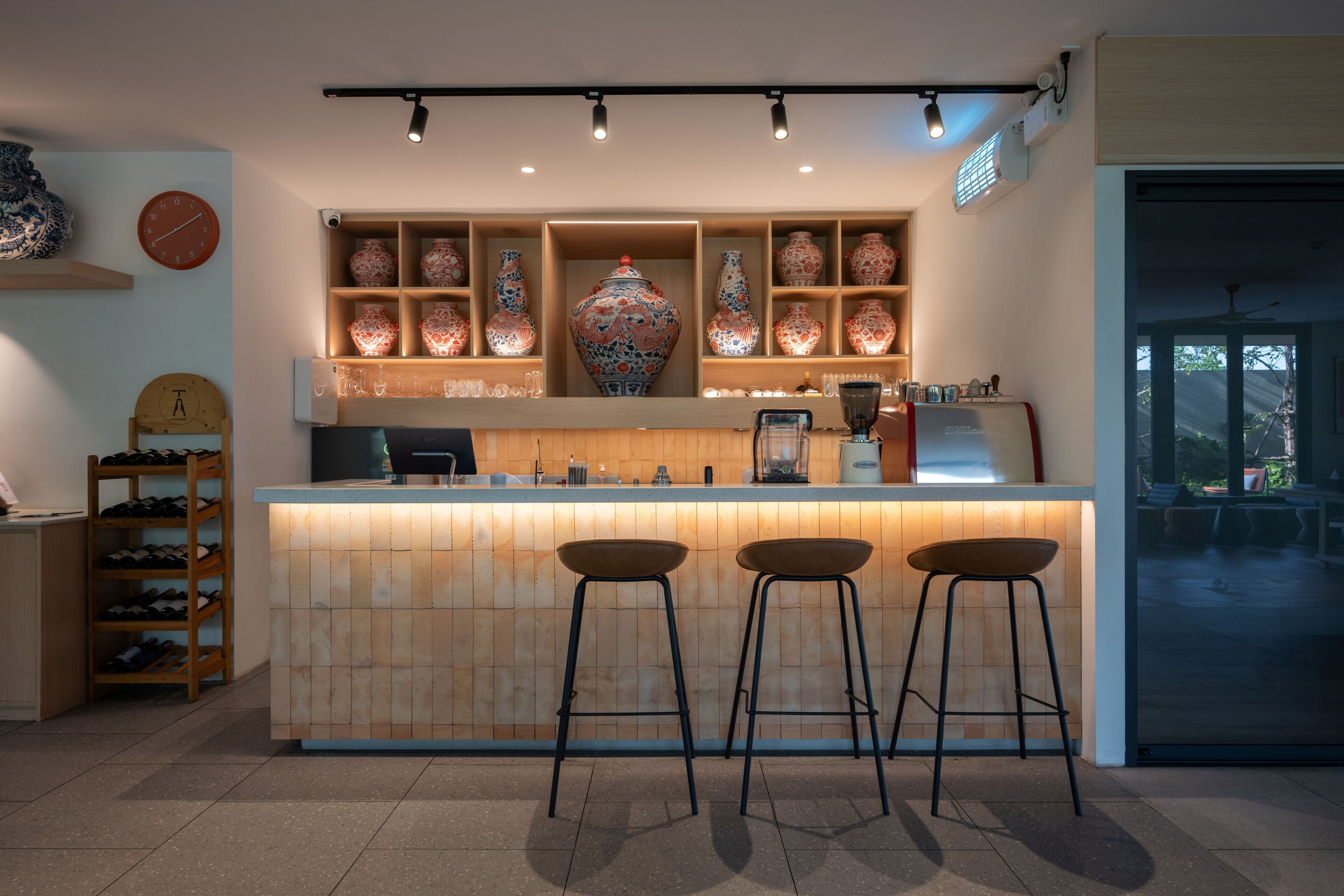 A bar area with stools, shelves with decorative vases, a clock, and a counter with a coffee machine.