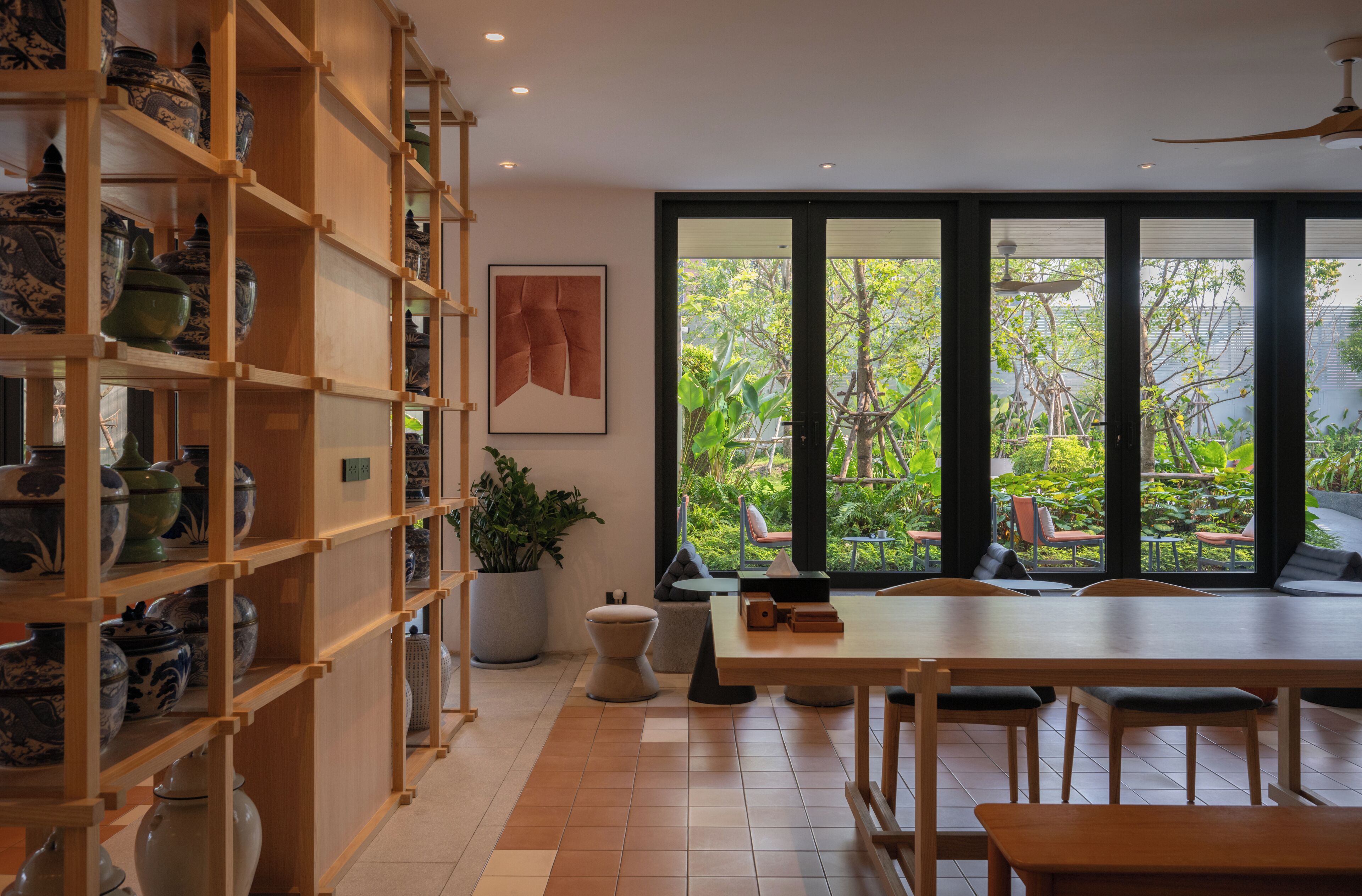 A dining area with a wooden table and chairs, a potted plant, and a view of greenery through large windows.