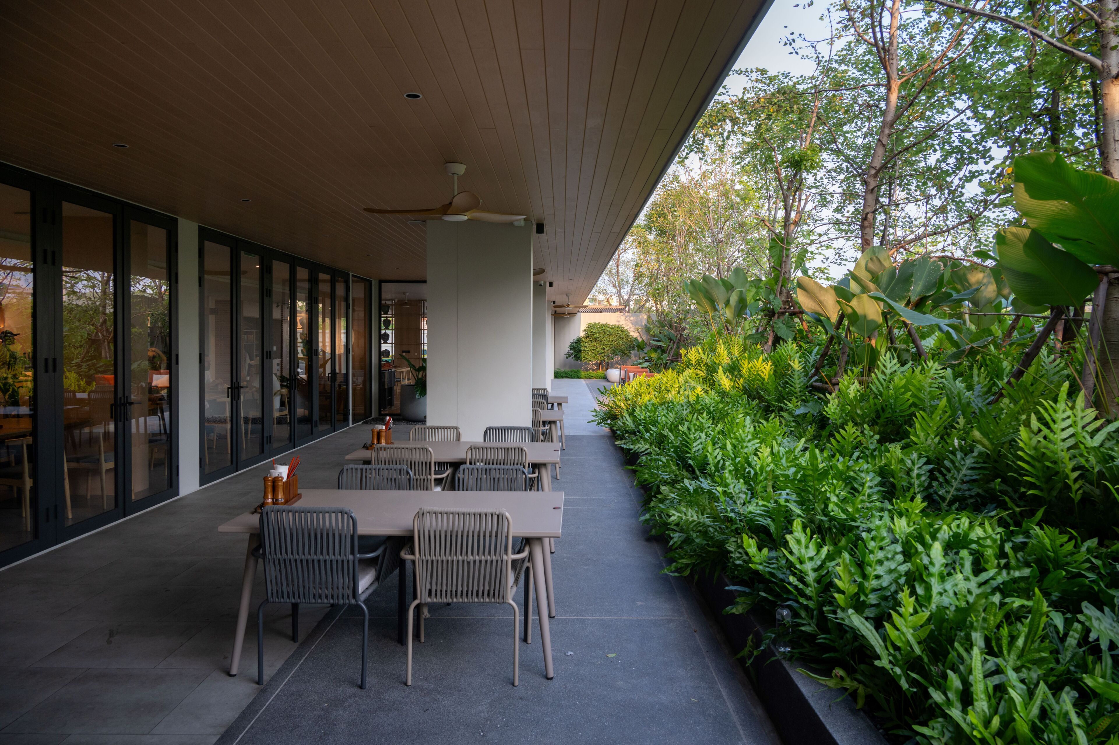 An outdoor dining area with tables and chairs, surrounded by lush greenery and a wooden ceiling.