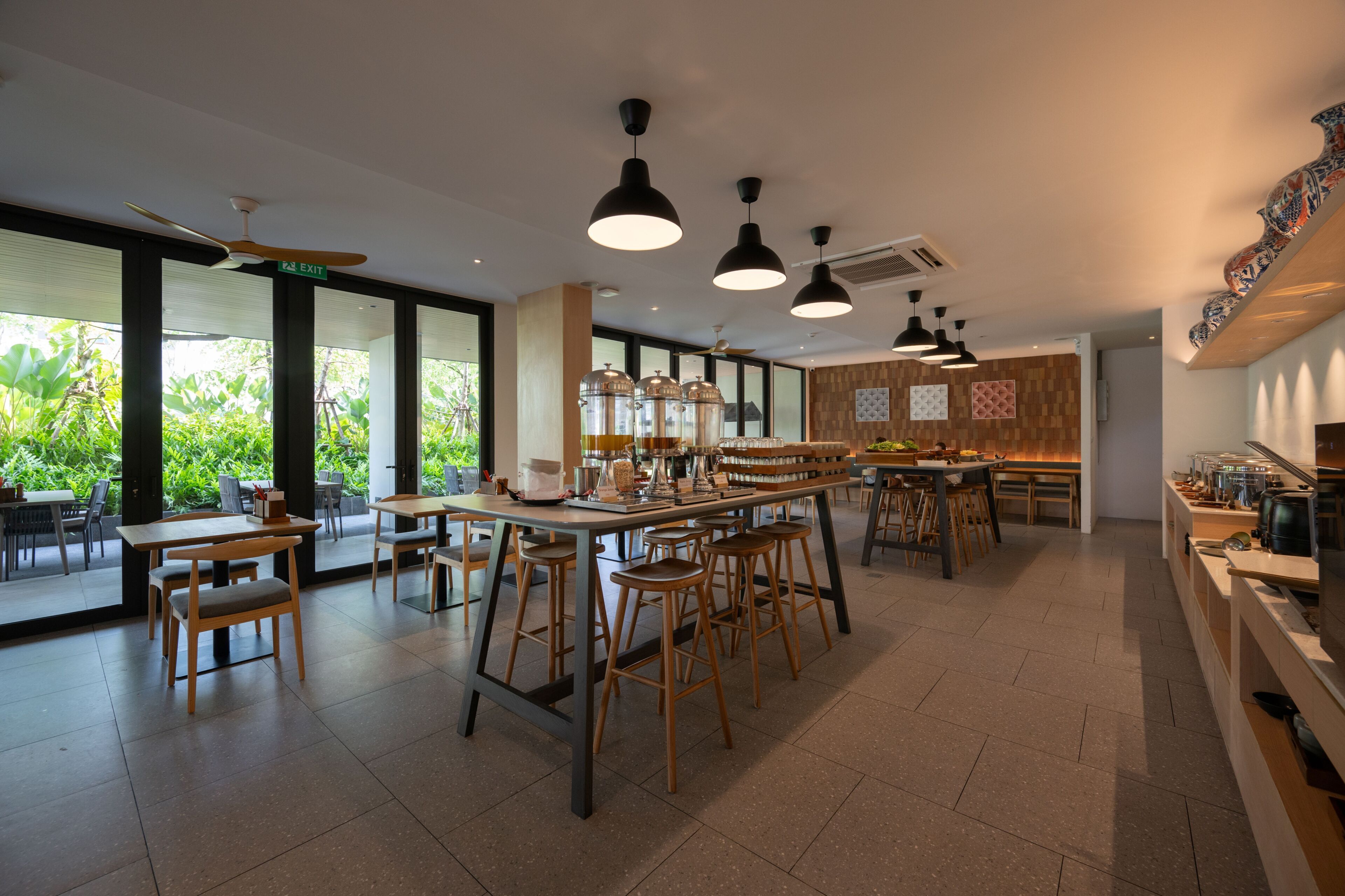 Un café spacieux, doté de nombreuses tables et chaises, d’un bar central et de grandes fenêtres donnant sur un espace verdoyant.