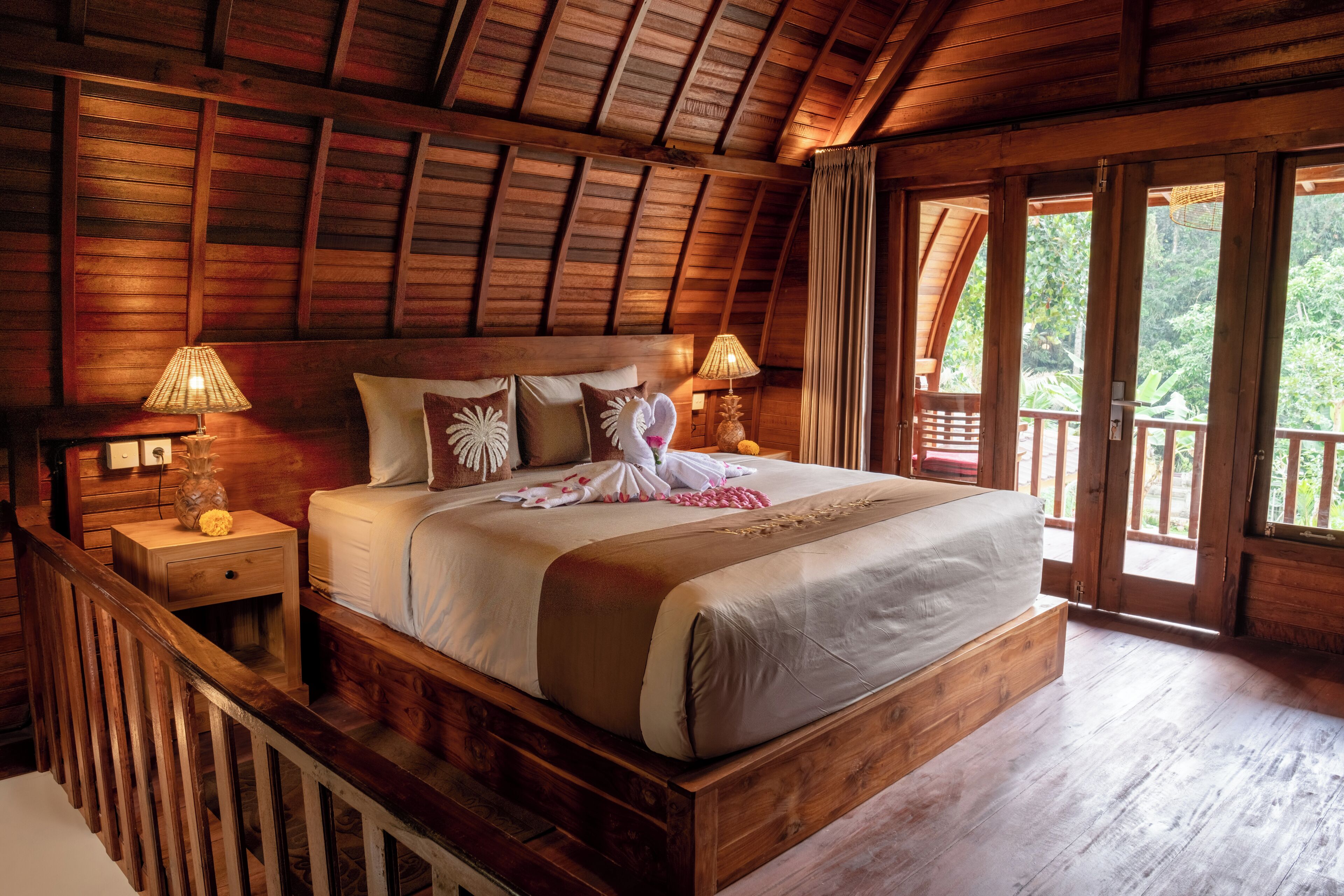 A wooden-framed bedroom with a large bed, two bedside tables with lamps, and a view of the outdoors through a sliding door.
