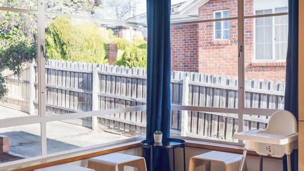 Una habitación con piso de madera, una ventana que da a una casa de ladrillo y una cerca de madera, una mesa con sillas y una lámpara colgante.