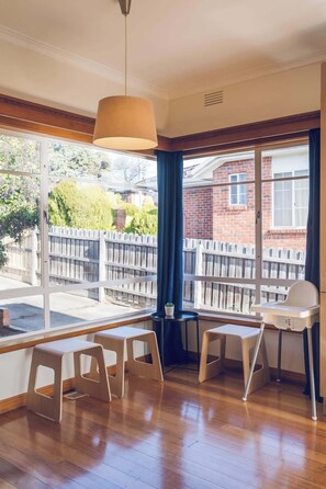 Une pièce avec un plancher en bois, une fenêtre donnant sur une maison en briques et une clôture en bois, une table avec des chaises et un luminaire suspendu.
