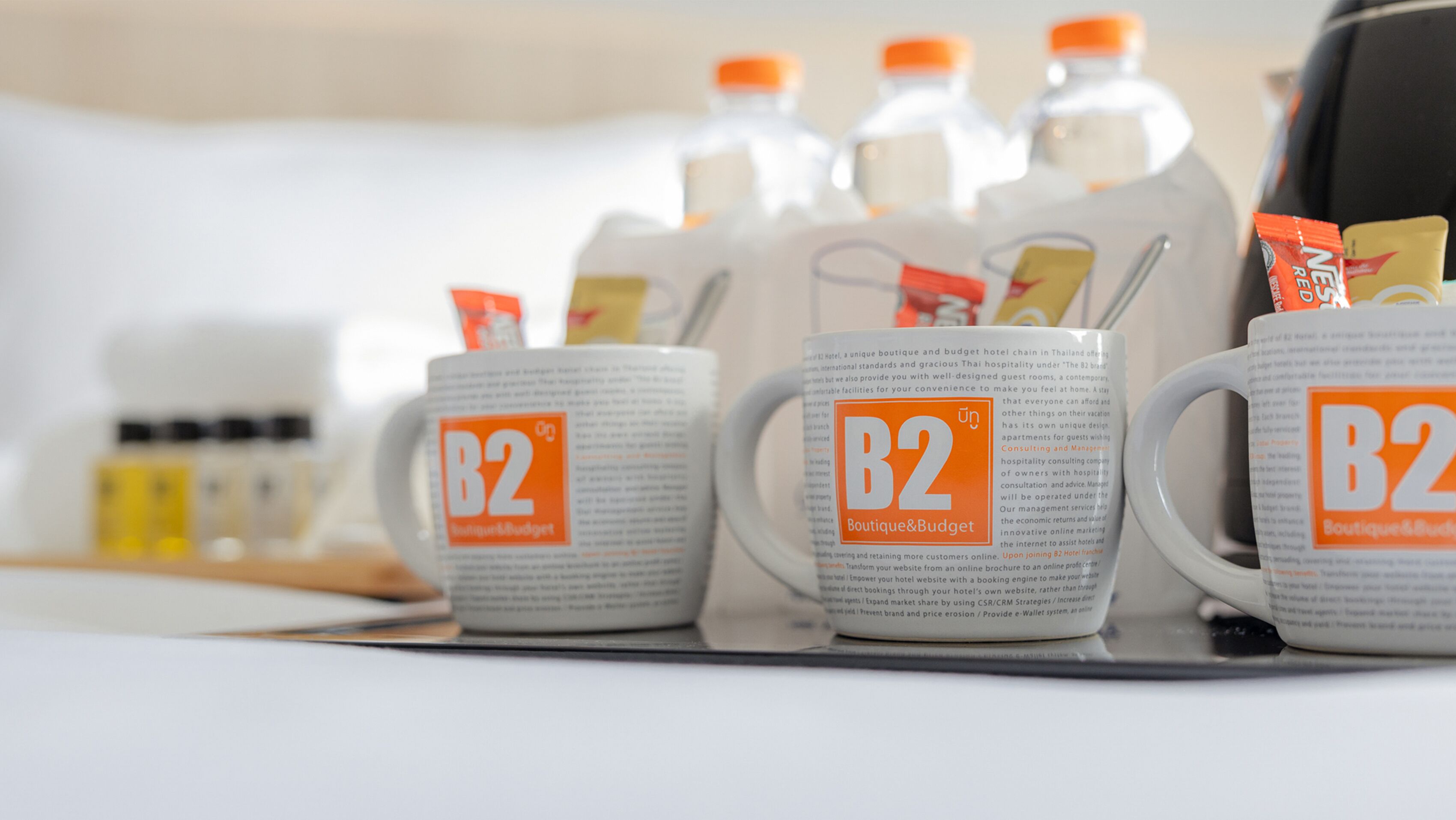 Trois tasses à l’effigie de la marque B2 sont posées sur une table, à côté d’un carton de lait et d’une boîte de thé.