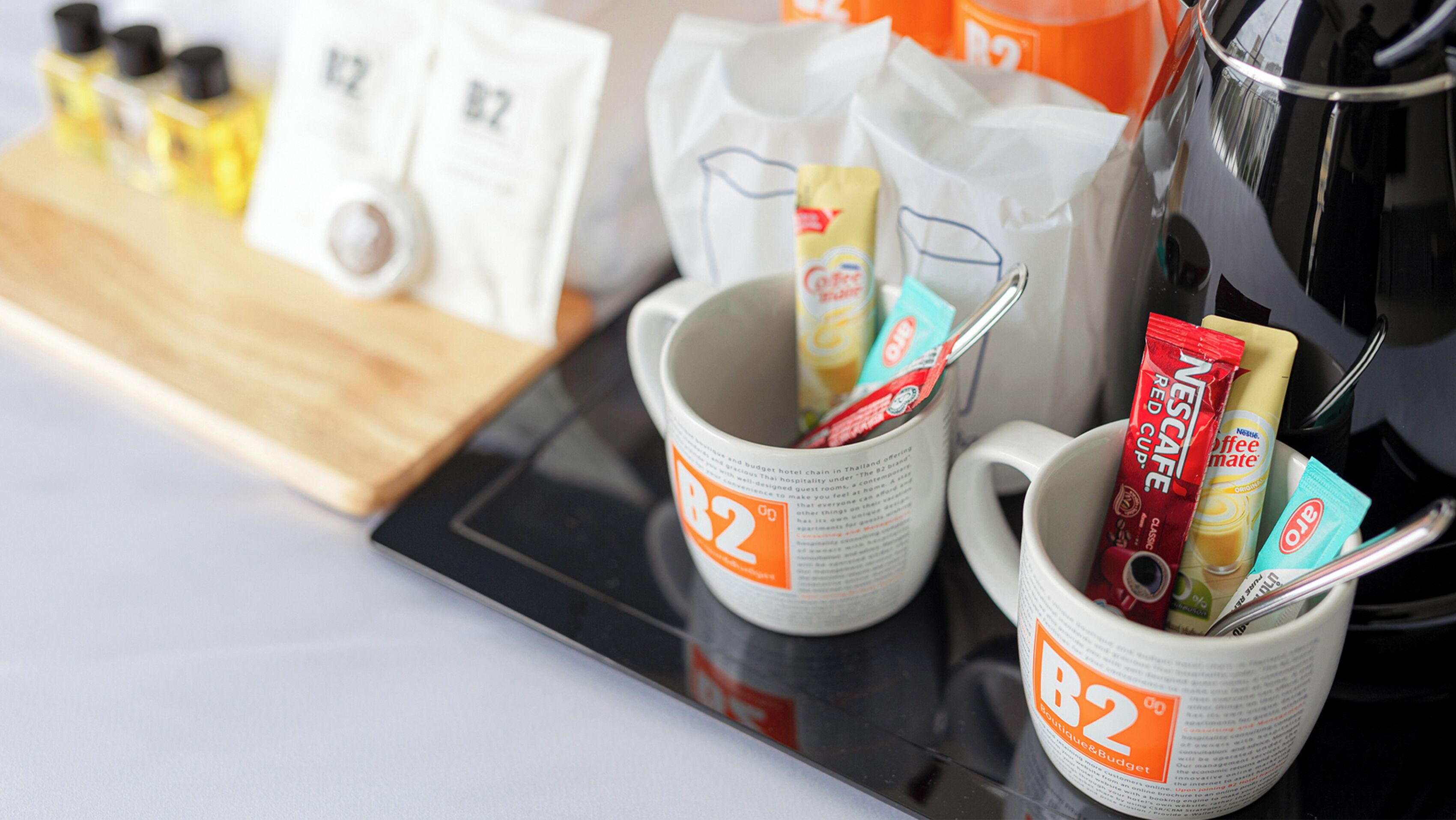 Two white mugs with B2 branding, filled with coffee and tea packets, and a coffee pot on a table.