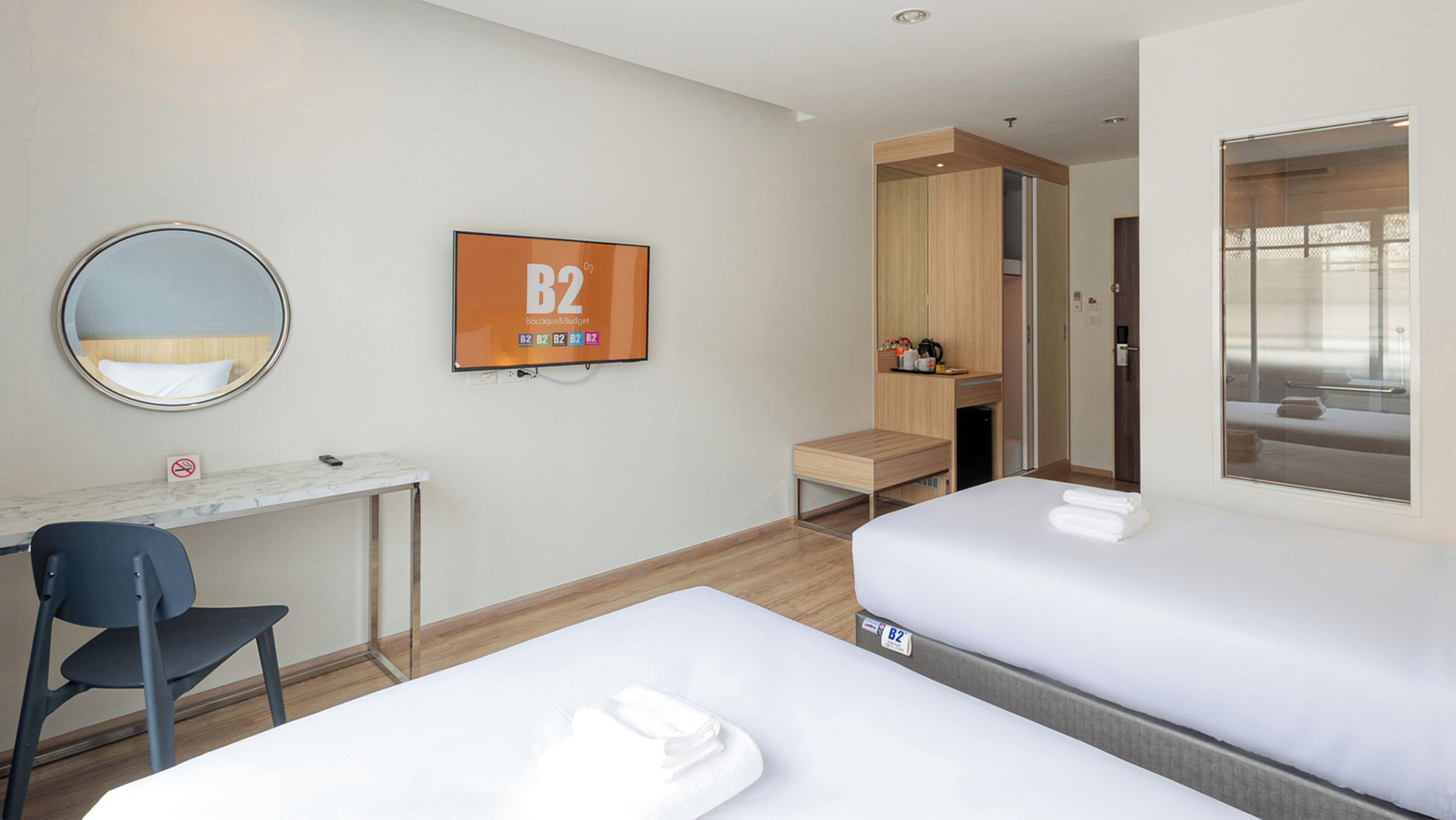 Une chambre d’hôtel avec deux lits, un bureau, une chaise, un miroir, un panneau encadré et une fenêtre.