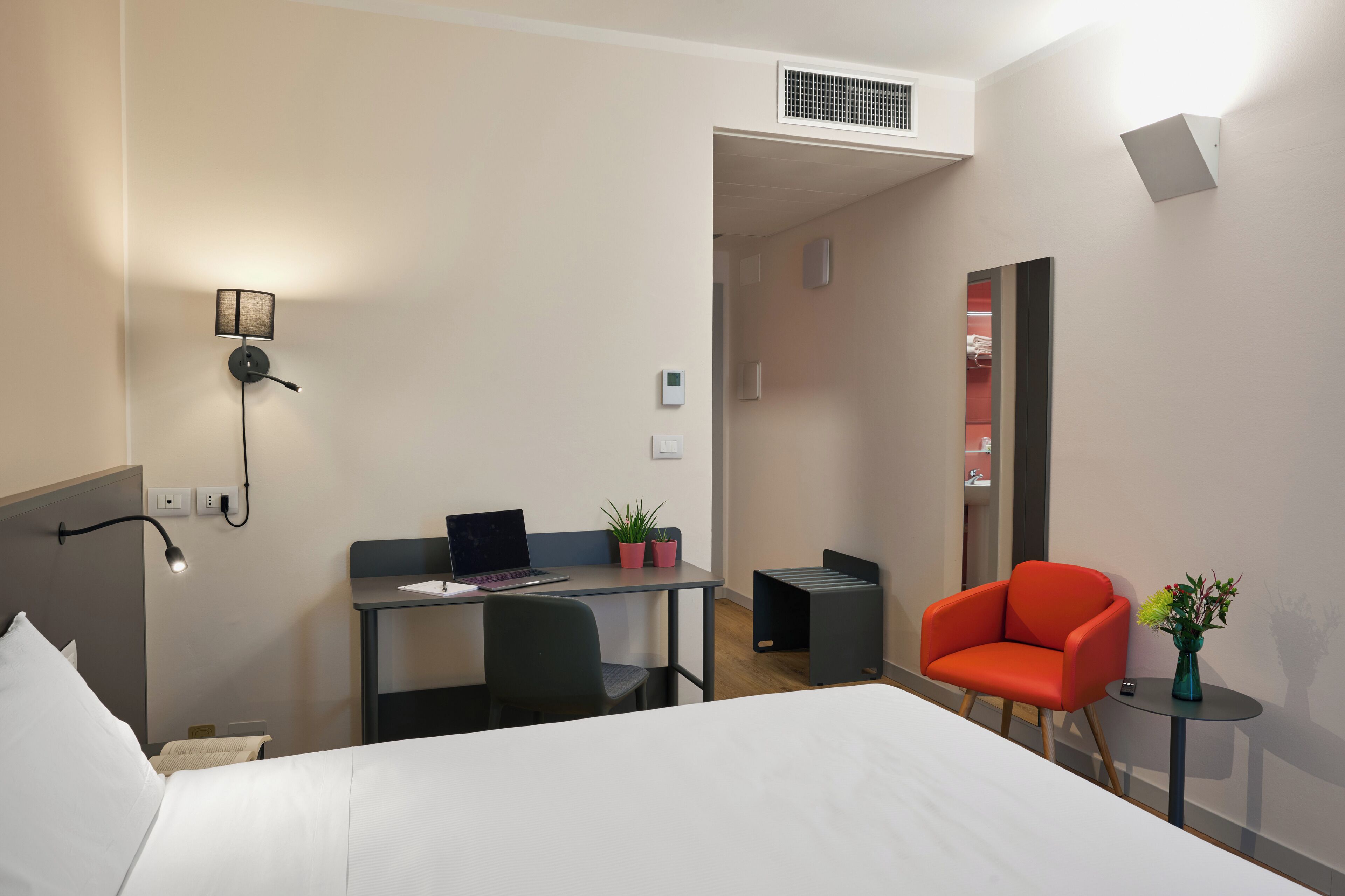 A hotel room with a bed, a desk with a laptop, a chair, a small table with a vase, and a mirror.