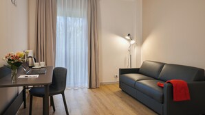 A modern living room with a sofa, a dining table with a laptop, a coffee maker, and a vase of flowers. - Zenit Park Hotel (Rivalta di Torino)
