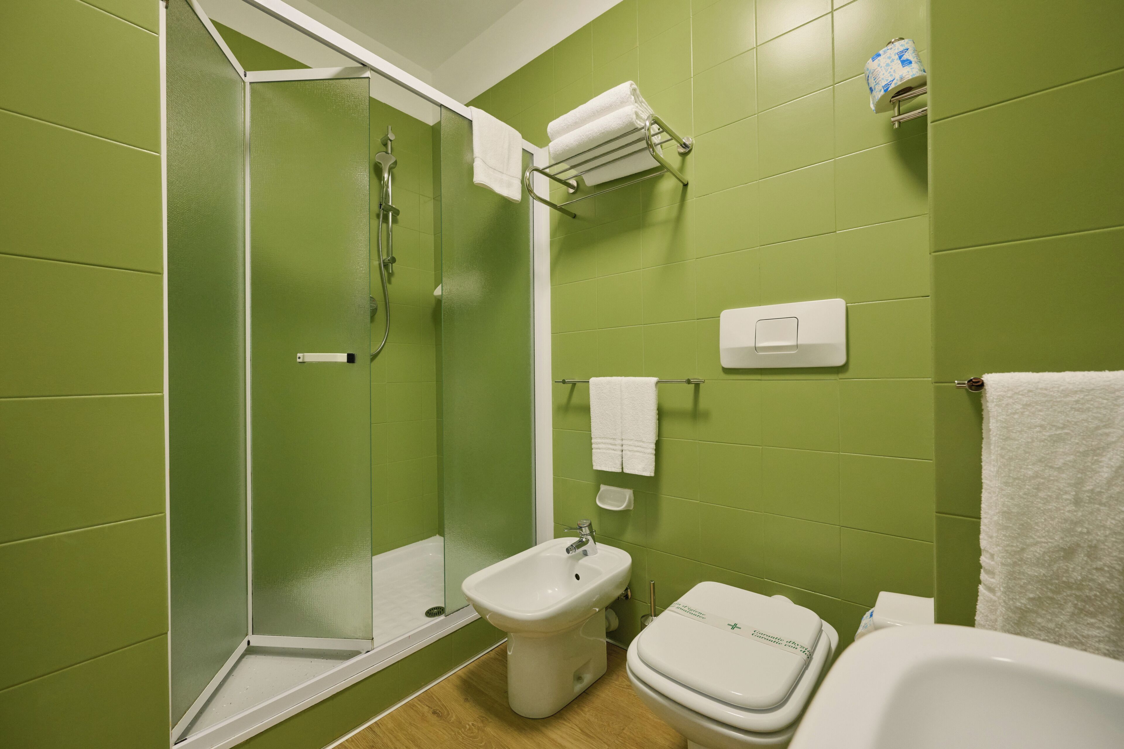 Un baño con paredes verdes, cabina de ducha de vidrio, lavamanos blanco, inodoro y toallero.