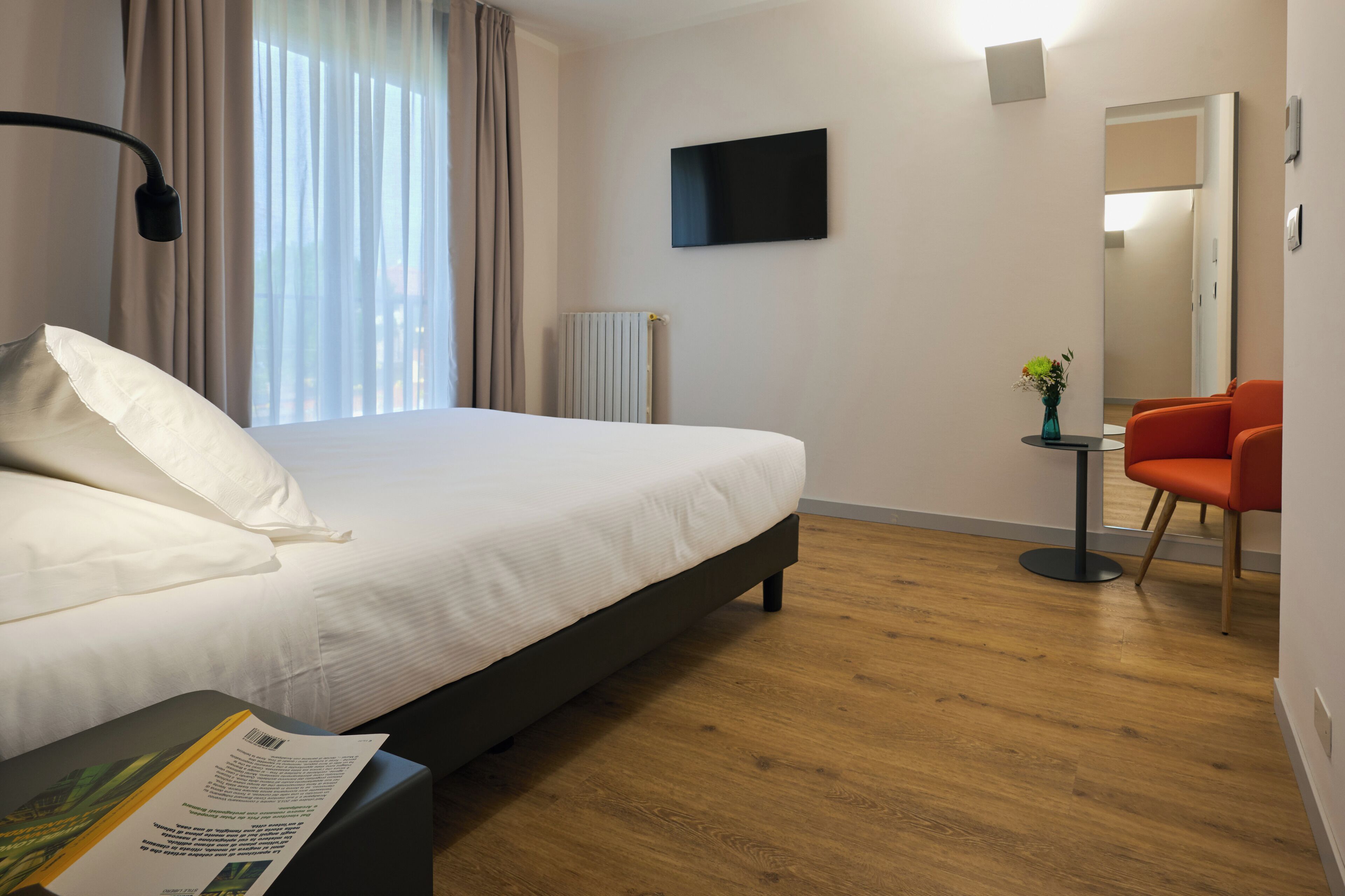 A hotel room with a bed, a desk with a chair, a TV, a mirror, and a small table with a vase.