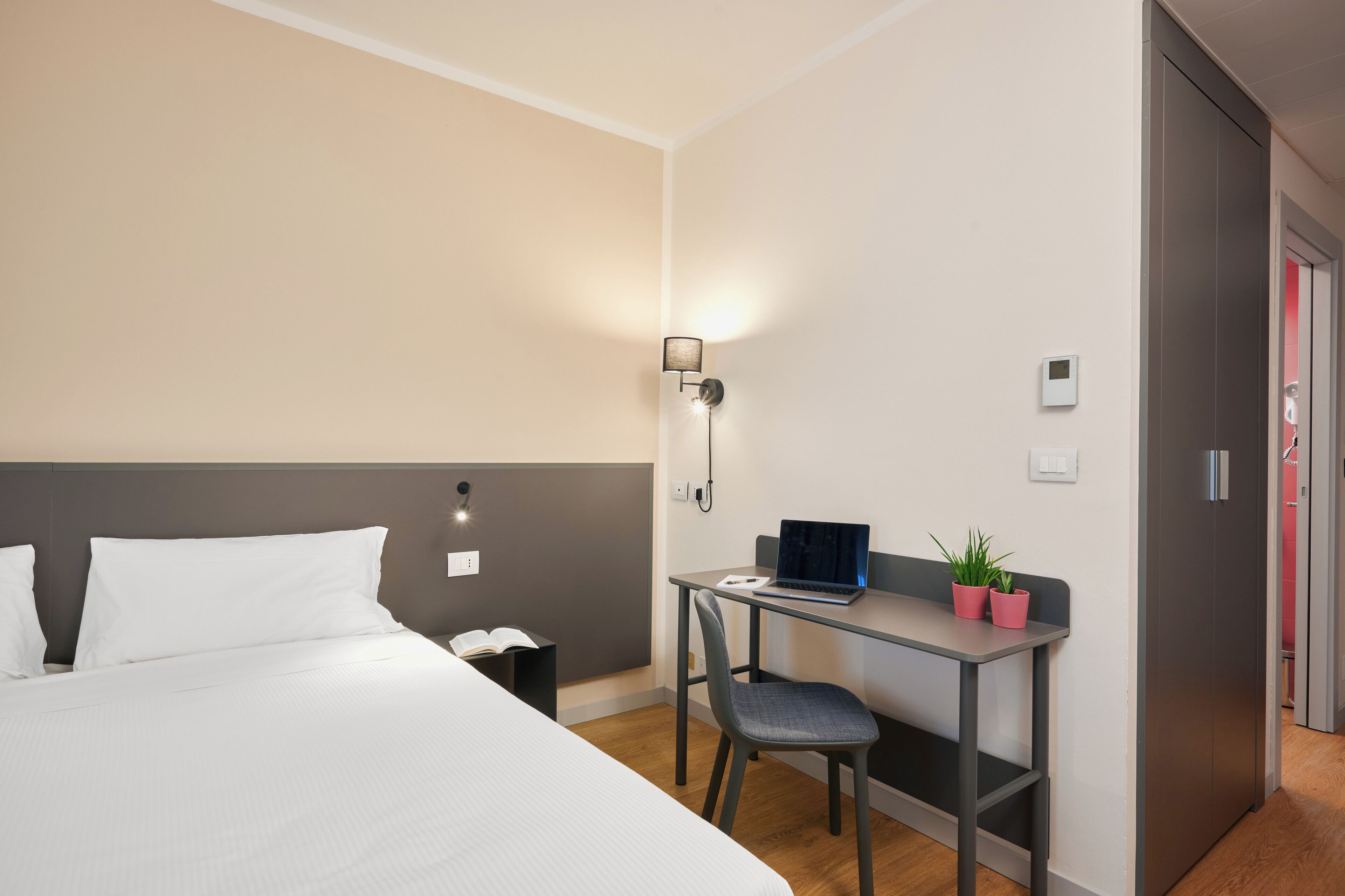 Una habitación de hotel con cama, escritorio con una computadora portátil, silla, pequeña mesa con una planta y mesita de noche.