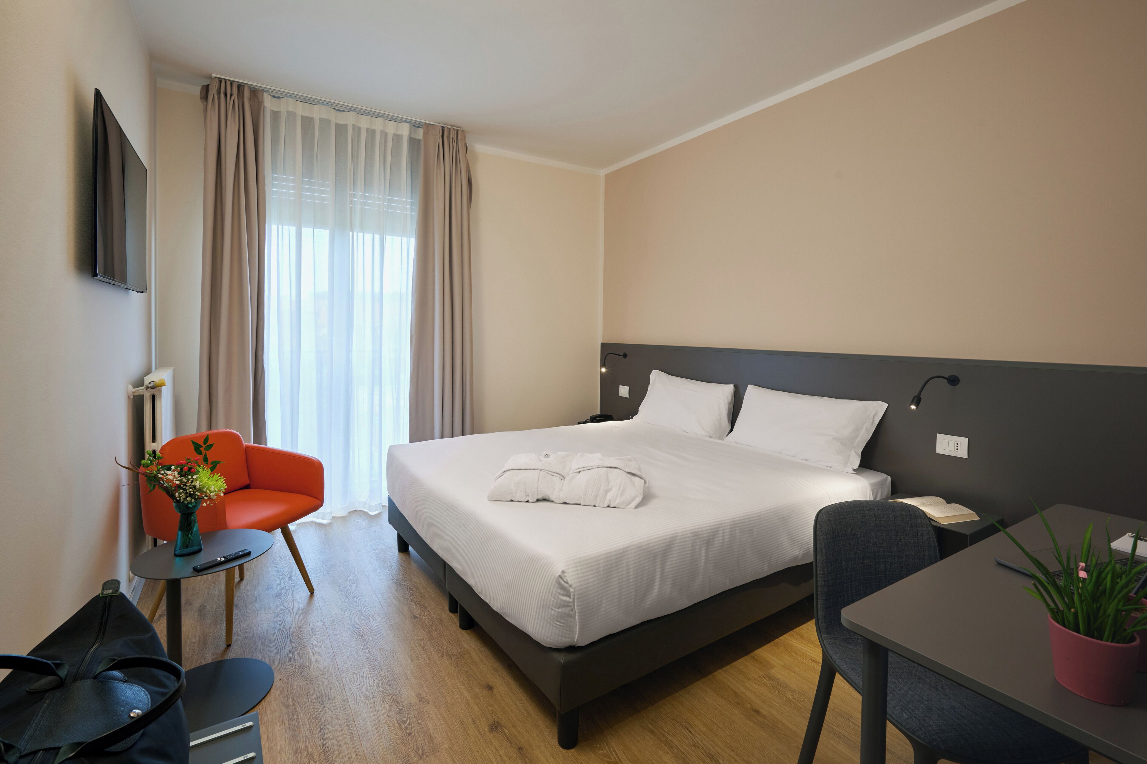 A neatly arranged hotel room with a bed, a desk, a chair, a small table, a plant, and a vase with flowers.