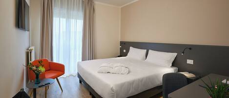 A neatly arranged hotel room with a bed, a desk, a chair, a small table, a plant, and a vase with flowers.