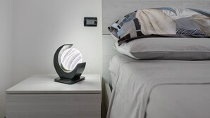 A bedside table with a modern lamp, a bed with patterned bedding, and a small wall-mounted control panel.