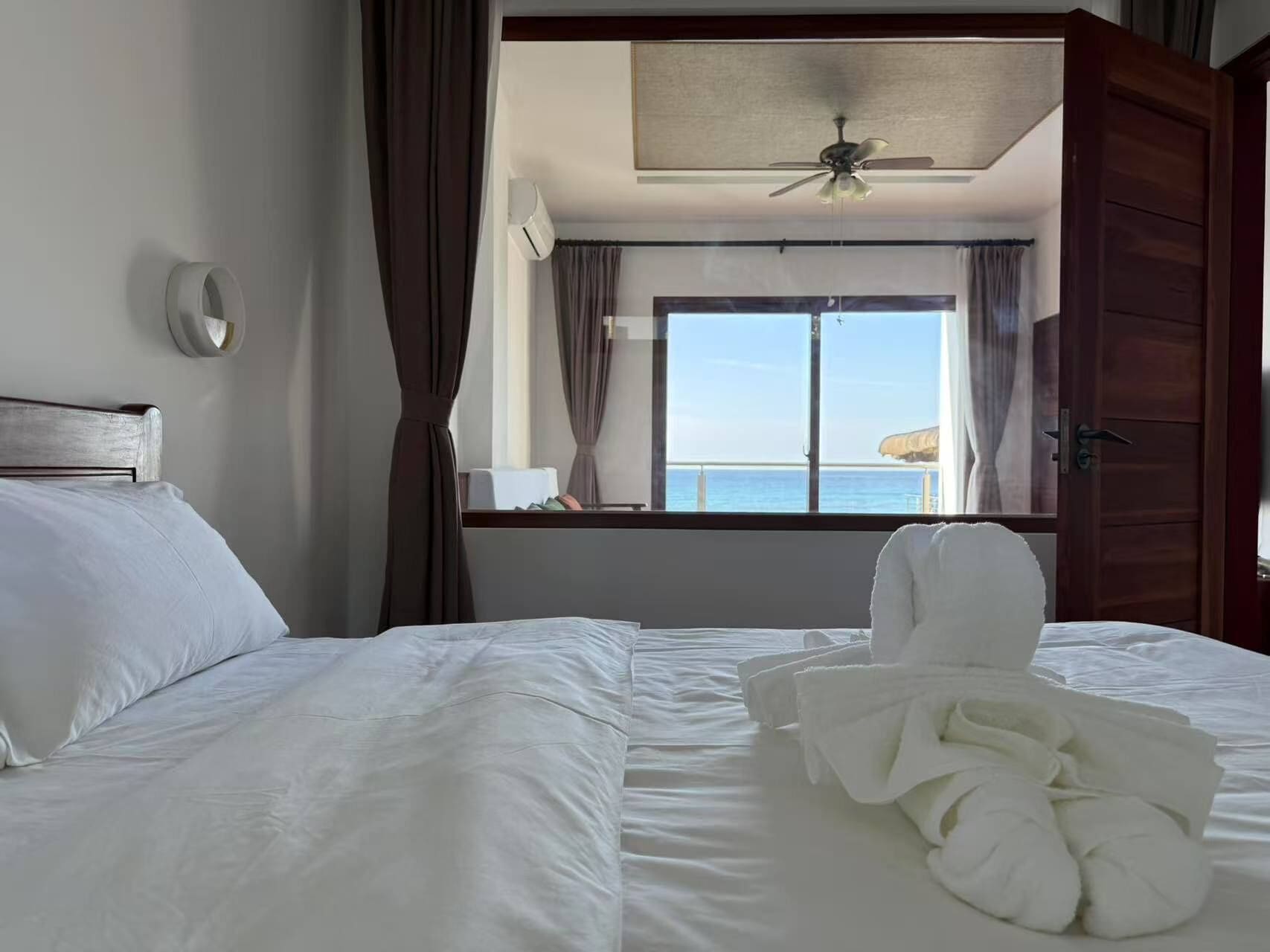A hotel room with a bed, a ceiling fan, a window with a view of the sea, and a door.
