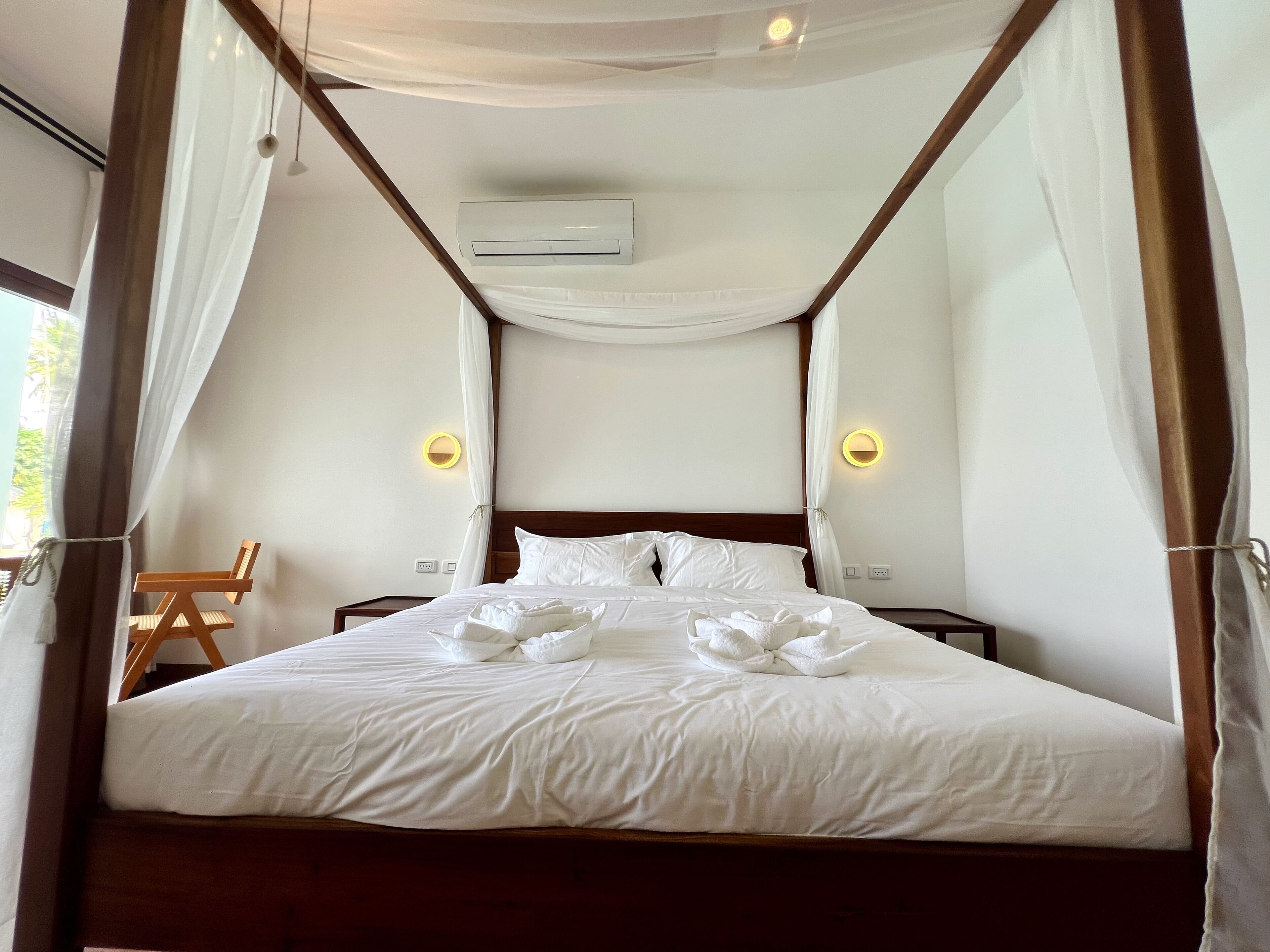 A four-poster bed with white linens and a canopy, two bedside lamps, and an air conditioning unit on the wall.
