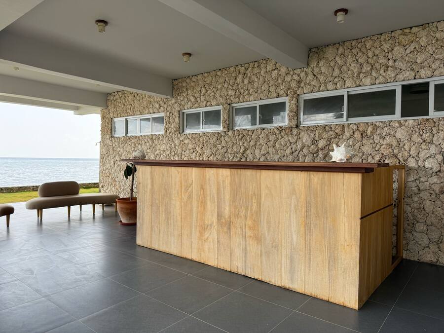 Zona de recepción con pared de piedra, mostrador de madera y asientos junto a la ventana con vista al mar.