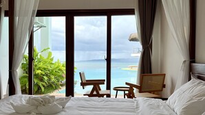 A hotel room with a large bed, two chairs, a small table, and a view of the sea through large windows.