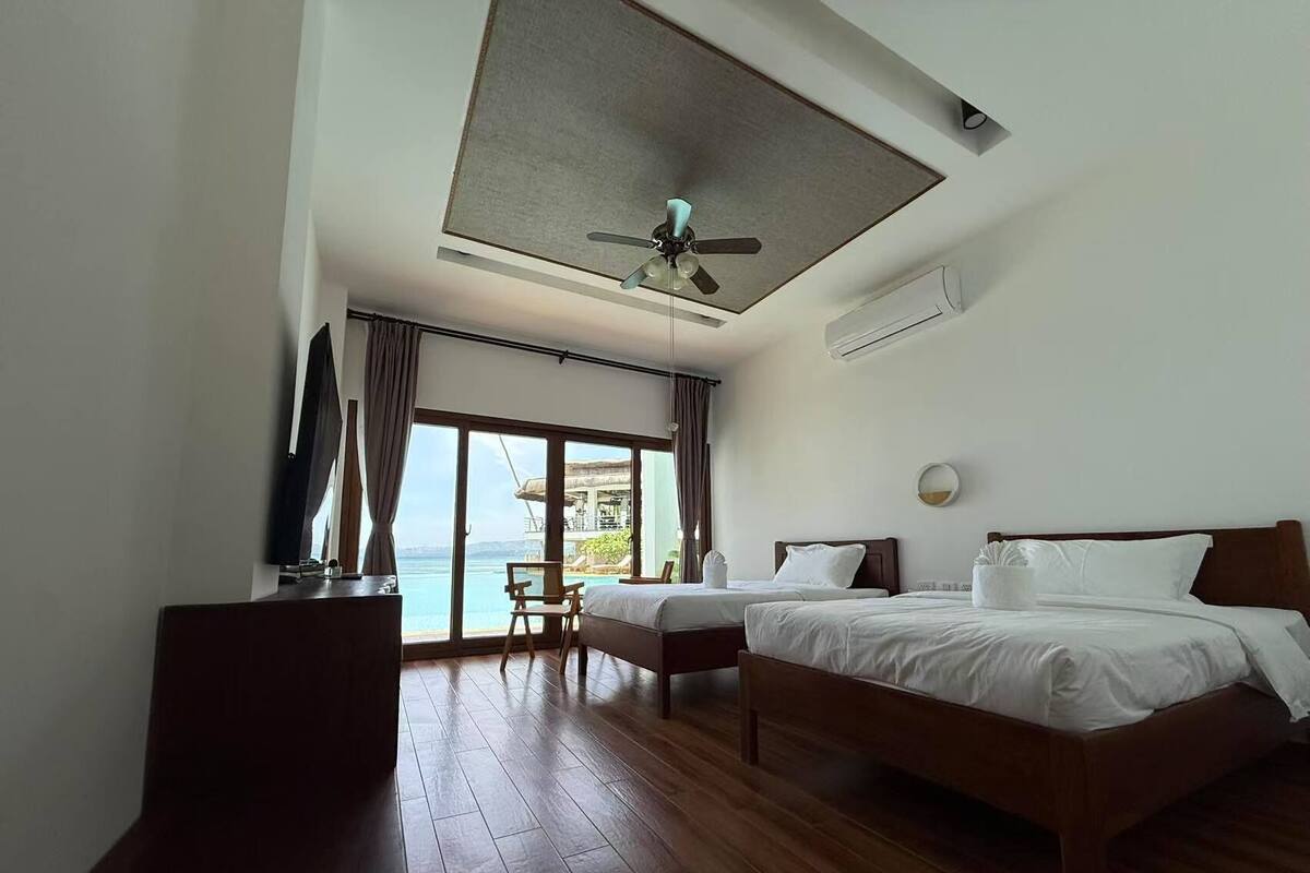 A hotel room with a large bed, a smaller bed, a desk, and a chair. There is a ceiling fan and an air conditioning unit. The room has large windows with a view of the ocean.